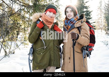 Horizontal medium portrait of active young Caucasian man and woman wearing backpacks posing on camera while hiking on winter day Stock Photo