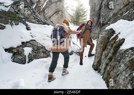 Young man holding hand of his girlfriends helping her to climb up while hiking in mountains on winter day Stock Photo
