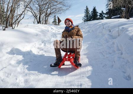 Low angle of cheerful young Caucasian man spending winter day outdoors sledding down the hill Stock Photo