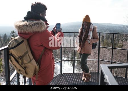 Young Black male tourist standing at observation deck on mountain top taking photos of his beautiful girlfriend Stock Photo
