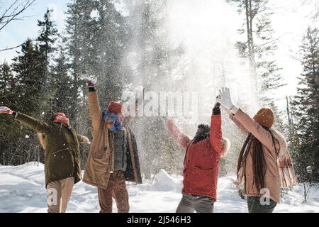 Group of young playful men and women enjoying spending time outdoors on winter day throwing snow in the air Stock Photo