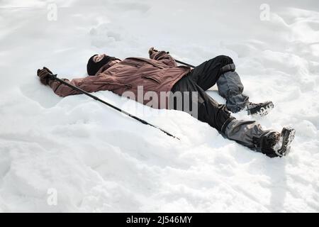 High angle shot of young guy wearing balaclava holding trekking poles relaxing on snowdrift on sunny winter day Stock Photo