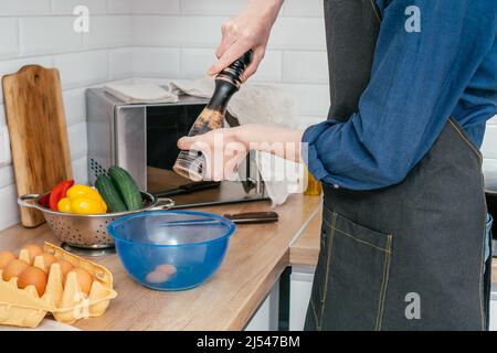 Side view of unrecognizable man peppering two eggs with wooden spice grinder in blue plastic bowl on table near steel colander with red, yellow bell p Stock Photo
