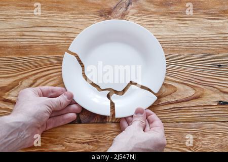 Top view on broken white plate and human hands holding two fragments on wooden background. Stock Photo