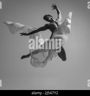 Flight. Black and white portrait of graceful muscled male ballet dancer dancing with fabric, cloth isolated on grey studio background. Grace, art Stock Photo