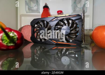 An overheated video card for mining or gaming on the refrigerator shelf with vegetables . Creative CPU cooling. Cryptocurrency Mining Stock Photo