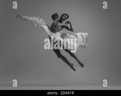 Black and white portrait of graceful muscled male ballet dancer dancing with fabric, cloth isolated on grey studio background. Grace, art, beauty Stock Photo