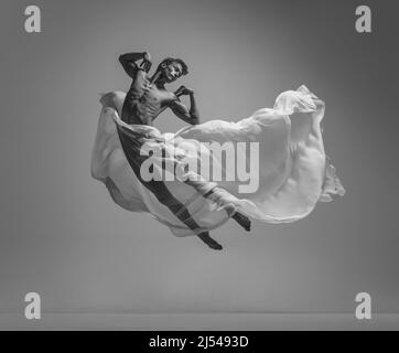 Solo. Black and white portrait of graceful muscled male ballet dancer dancing with fabric, cloth isolated on grey studio background. Grace, art Stock Photo