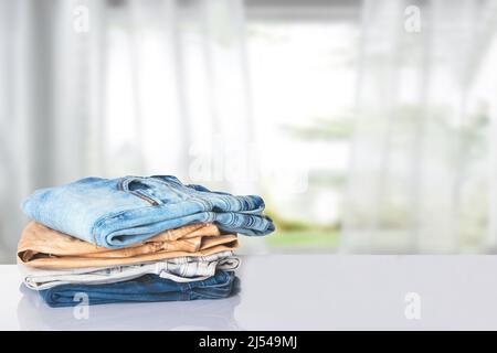 Stack of jeans. Closeup of a pile of colorful female denim pants on a bright table against abstract blurred curtain background. Copy space. Stock Photo