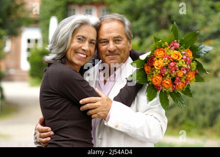 elderly couple in the park, man lovingly hugs her after handing her a bouquet of flowers Stock Photo