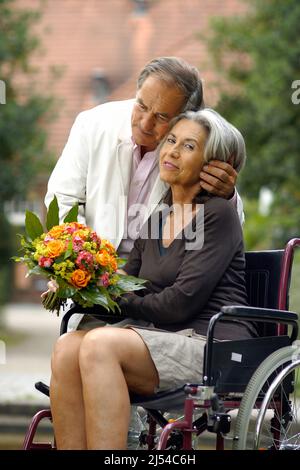 elderly couple in the nature, the woman is sitting in a wheelchair with a bouquet of flowers and the man is hugging her affectionately Stock Photo