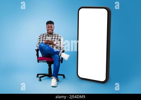 Black Man Using Cellphone Sitting Near Huge Phone, Blue Background Stock Photo