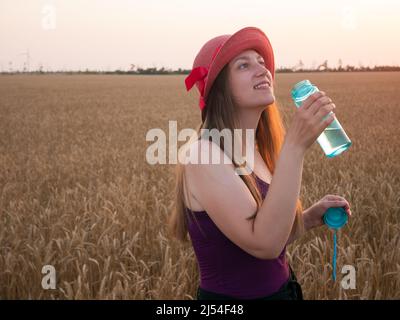 Young girl in hat holds a bottle of water in hand and drinks water in wheat field. Weight loss, summer active lifestyle body care. Rehydration body in Stock Photo