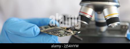 Engineer in glove holds microcircuit against background of microscope Stock Photo
