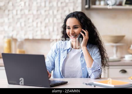 Portrait of happy latin lady working from home, talking on cellphone and using laptop, sitting at table in kitchen Stock Photo
