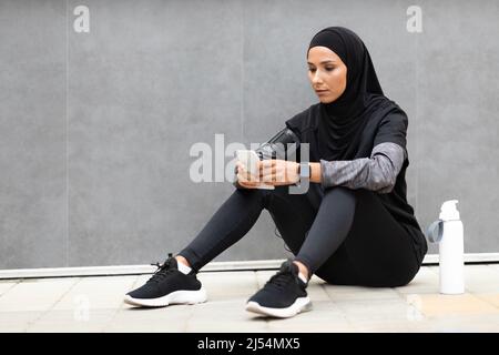 Confident young arab lady athlete in hijab, sportswear with phone and water bottle, sits on gray wall background