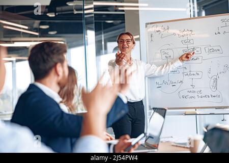 Experienced businesswoman by whiteboard explaining situation on the market to young managers Stock Photo