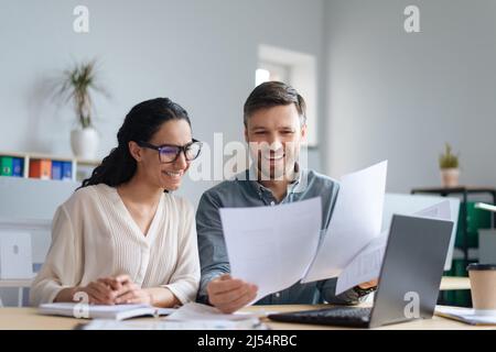 Cheerful male and female company workers looking through documents, collaborating, using laptop at modern office Stock Photo
