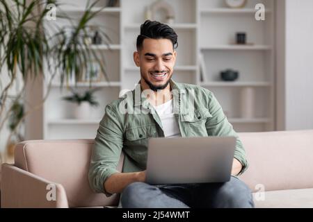 Happy attractive millennial arabic male with beard in casual sits on sofa, typing on laptop in living room Stock Photo