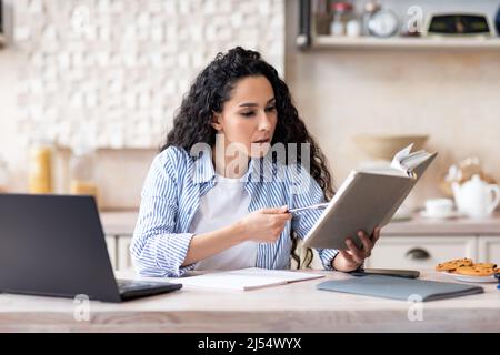 Concentrated lady sitting in kitchen, using laptop and reading book, studying remotely from home Stock Photo