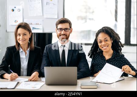 Successful multiracial influential business people, elegant creative colleagues sitting in modern office, working together on project, using laptop, analyze financial documents, lookin at camera,smile Stock Photo