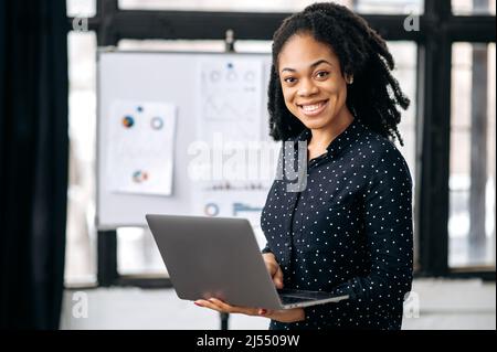 Portrait pretty African American business woman, financial manager or mentor, broker, marketer, stand in office, against a white board with charts, holds an open laptop in hands, looks at camera,smile Stock Photo