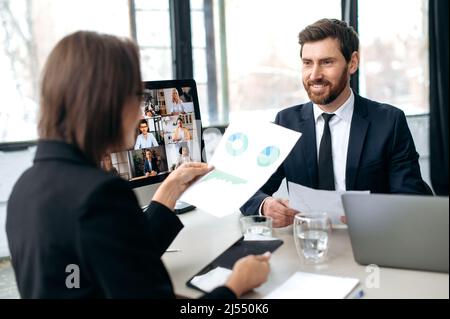 Two influential successful business colleagues, working together in modern office, study financial documents, talking with partners by online video conference by computer, discuss business strategy Stock Photo