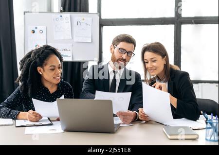 Business partners working together on a project. Multiracial colleagues are brainstorming on strategy together, sitting in modern office, studying documents, using laptop, discussing, smiling Stock Photo