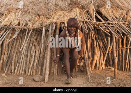 Pregnant Hamar woman with necklaces made of Cowry shells coming out of her wooden hut, Omo river valley, Southern Ethiopia Stock Photo