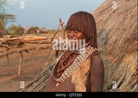 Pregnant Hamar woman with necklaces made of Cowry shells in front of her wooden hut, Omo river valley, Southern Ethiopia Stock Photo