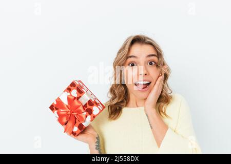 Amazed happy young woman received an unexpected gift, holds red box in her hand and surprised looking at camera, white studio background Stock Photo