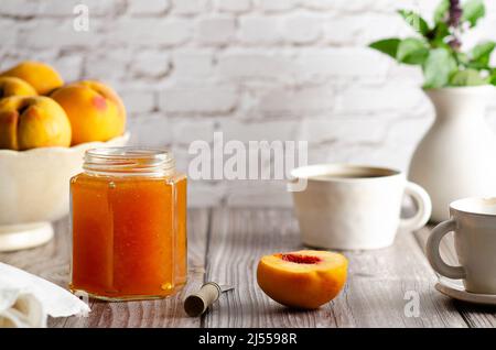 Homemade peach jam in a jar with some fresh peaches, two cups of coffee and a vase with green leaves, on a wooden table background. Stock Photo