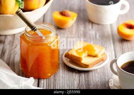 Homemade peach jam in a jar with some fresh peaches, two cups of coffee and some toasts, on a wooden table background. Stock Photo
