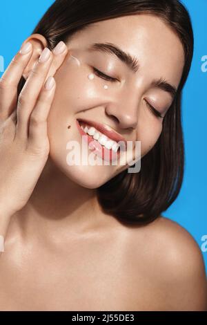 Close up of girl applies lotion, facial skin care cream product on face and smiles from pleasure, nourish face with spa cosmetic product, stands over Stock Photo
