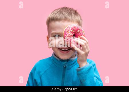 A cute funny boy in a blue sweater holds a bright pink donut near his eye and laughs on a pink background. Adorable cheerful kid is playing with a don Stock Photo