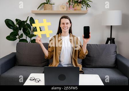 Smiling attractive young woman blogger sitting on comfortable sofa at home showing big yellow hashtag symbol and smartphone, happy looking at camera, Stock Photo