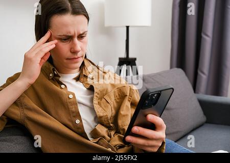 Sad female feeling upset reading bad news in mobile message on smartphone, frustrated young woman sitting on couch holds phone scared of threatening s Stock Photo