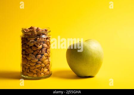 Peanuts in a glass jar and green apple on a yellow background Stock Photo