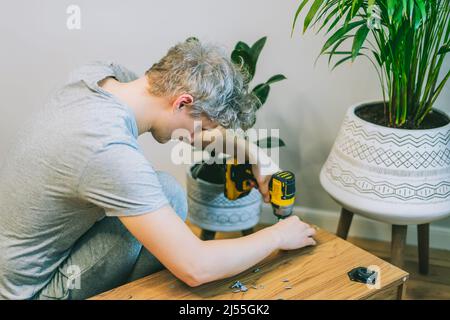 A young man with an electrical screwdriver assembles a tv stand console according to instructions in his new house. Man assembling furniture at home u Stock Photo