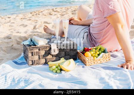 Summer beach picnic at sunset. Young man sitting on blanket having weekend picnic outdoor at seaside with fresh seasonal fruit, cheese and glasses of Stock Photo