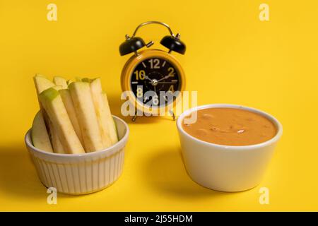 Sliced ​​apple and peanuts butter in cups with alarm clock in the background on a yellow background Stock Photo