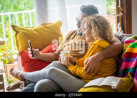Young adult caucasian couple enjoy life together at home doing video call at friends sitting on the sofa and enjoying wireless connection. Modern peop Stock Photo