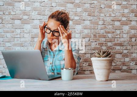 Woman working at the desk on laptop with headache. Concentrated and worried female businesswoman in front of a computer. Home work place with wall bri Stock Photo