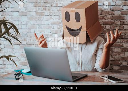 Happy modern online worker wearing smile carton box on head speaking in video call at the laptop. Internet job business and identity concept. Customer Stock Photo