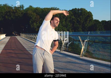 Handsome middle-aged Caucasian man, sportsman, athlete exercising outdoor, standing on a treadmill, warming up his body, stretching it before morning