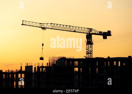 Silhouette of tower crane with cargo and workers on building scaffolding at sunrise. Housing construction, apartment block in city on dramatic sky Stock Photo