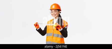 Pleased smiling asian female architect achieve good results, looking satisfied at tape measure after measuring layout at construction area, standing Stock Photo