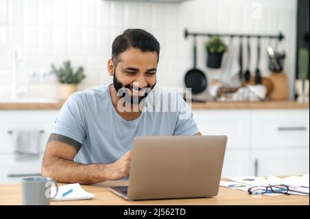 Positive handsome successful indian or arabian man, freelancer, designer, IT specialist, sitting at the table in the kitchen with a laptop, working online on a new project, smiling happily Stock Photo