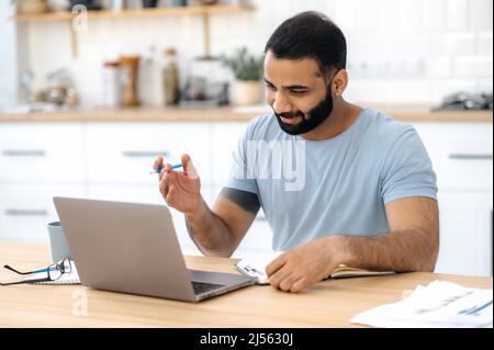 Positive modern Indian man, freelancer, designer or student, sitting at a kitchen at home, studying or working online uses laptop, smiling friendly, takes notes. E-learning, video conference concept Stock Photo