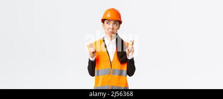Sad nervous and unconfident asian female construction manager in safety helmet, pouting and look in panic upper left corner, making wish, cross Stock Photo